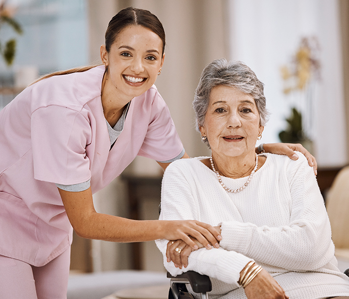 Smiling caregiver in pink scrubs assisting an elderly woman in a wheelchair, both looking at the camera in a bright, welcoming indoor setting