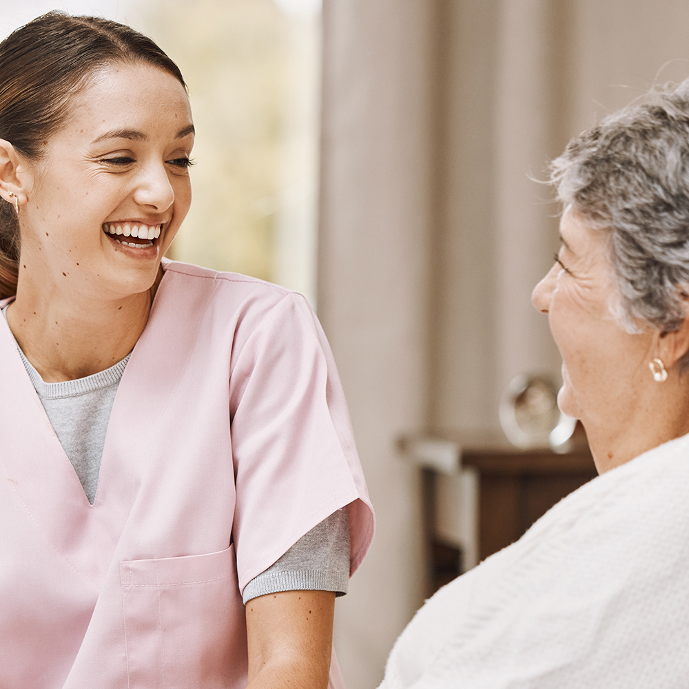 Caregiver in pink scrubs smiling and talking with an elderly woman in a home setting.